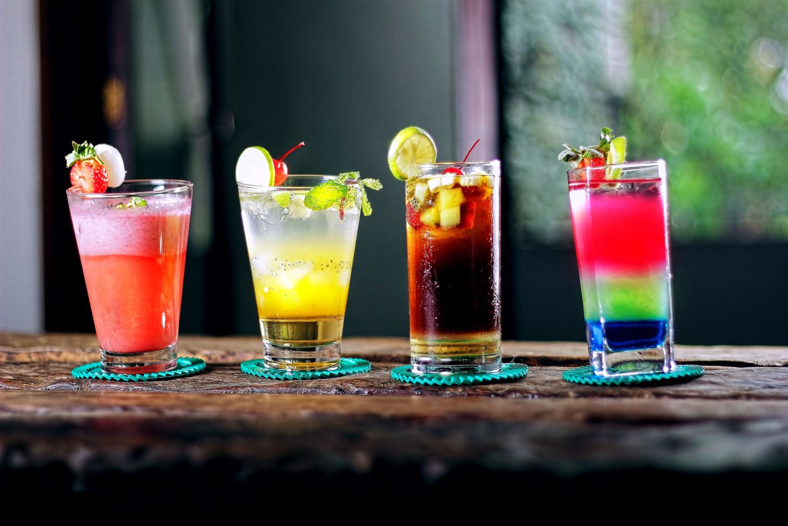 Four colorful cocktails with fresh fruit garnishes lined up on a rustic wooden table